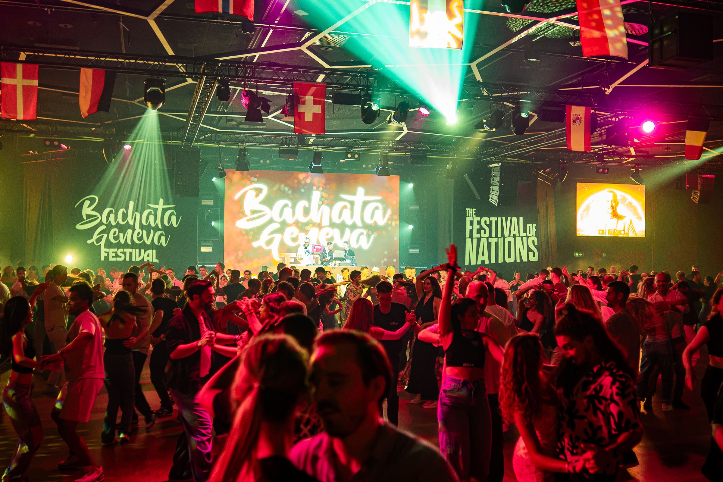 The main room of Bachata Geneva Festival 2026 at night, full of dancers under the flags